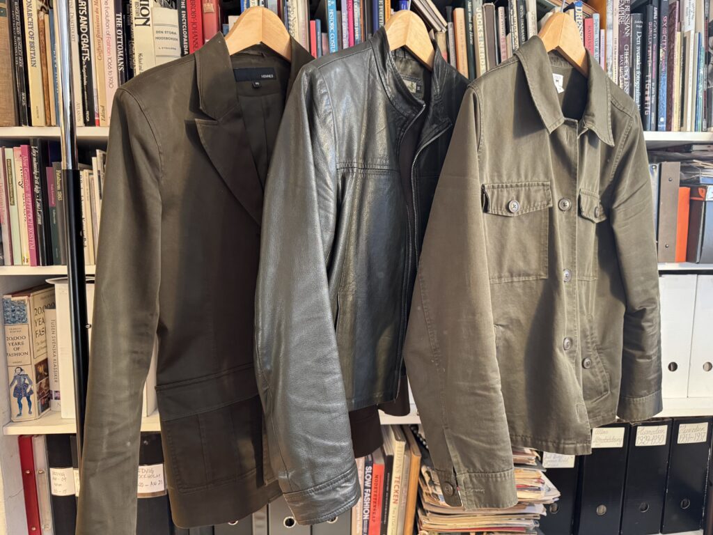 Tre gröna kavajer - en blazer, en skinnjacka och en button-up - hänger på trägalgar framför en bokhylla fylld med böcker och pärmar.