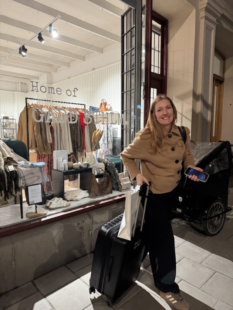 En kvinna i brun kappa ler när hon står utanför en upplyst butik med namnet "Home of GARDEROBSLIM". Hon håller i en resväska och en shoppingväska på en trottoar i en stad på natten. Kläder och accessoarer syns i skyltfönstret.