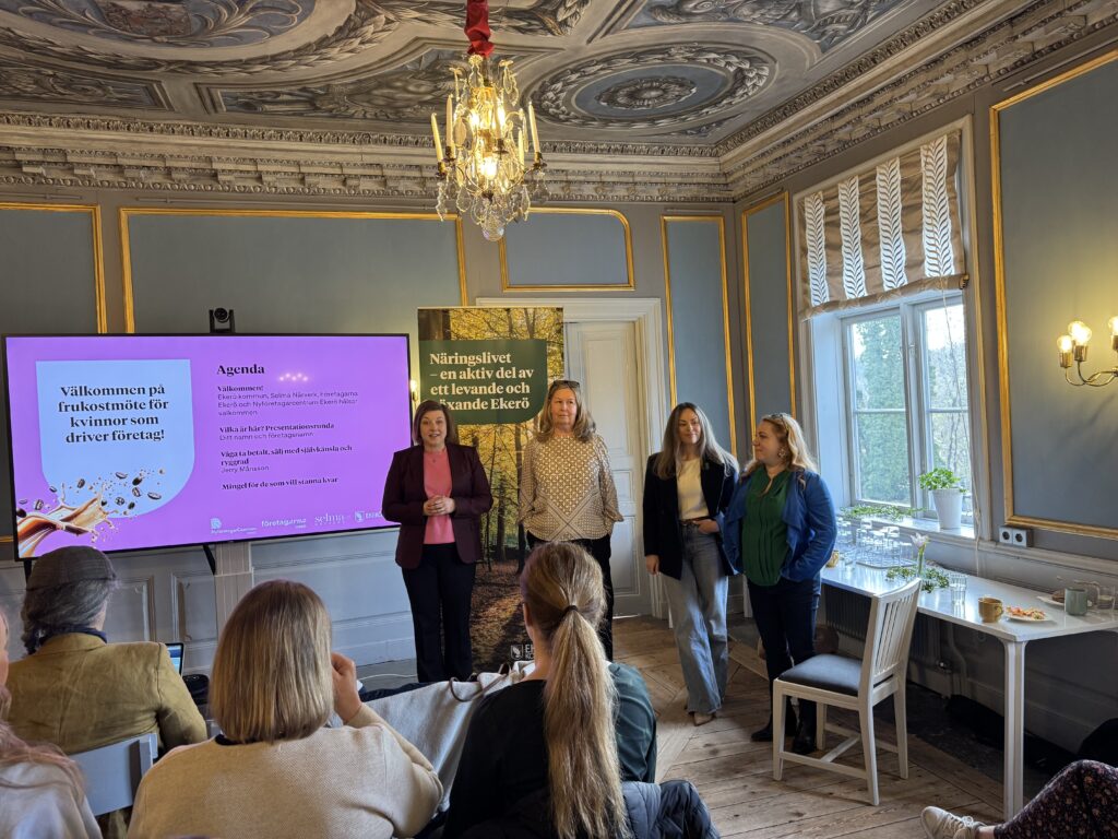 Fyra kvinnor står längst fram i ett elegant rum med utsmyckat tak och kristallkrona och presenterar sig för en publik. På en stor skärm visas en välkomsthälsning och en agenda på svenska för ett affärsevent för kvinnor.
