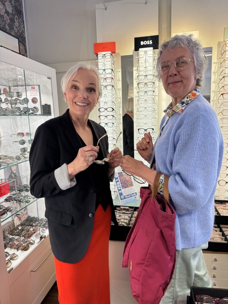 Två leende äldre kvinnor står i en glasögonbutik med varsitt par glasögon i handen. De är omgivna av montrar fyllda med glasögonbågar. Den ena kvinnan har en röd kjol och den andra bär en rosa väska.