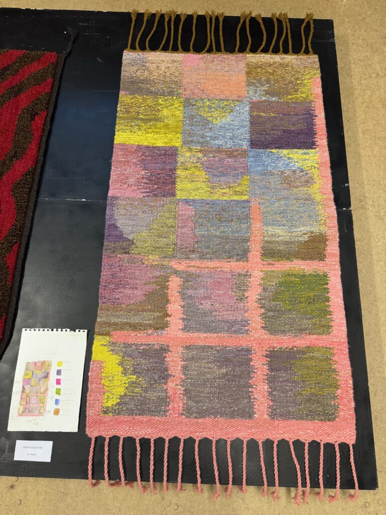 Rektangulär vävd textil med rutmönster i rosa, gula, lila och bruna toner. Rosa fransar finns i båda ändar. Ett färgreferenskort med en liten bild av textilen ligger till vänster.