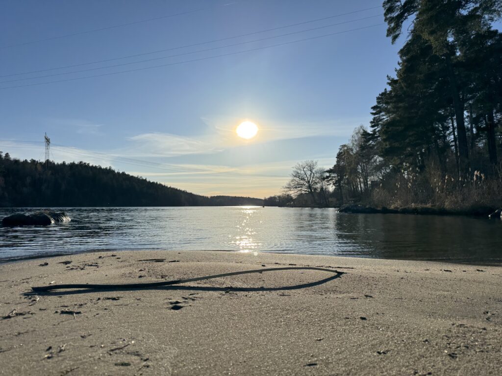 En sandstrand vid en lugn flod med träd på båda sidor, badande i solljus under en klarblå himmel. Solen reflekteras på vattenytan och i förgrunden ligger en pinne på sanden.