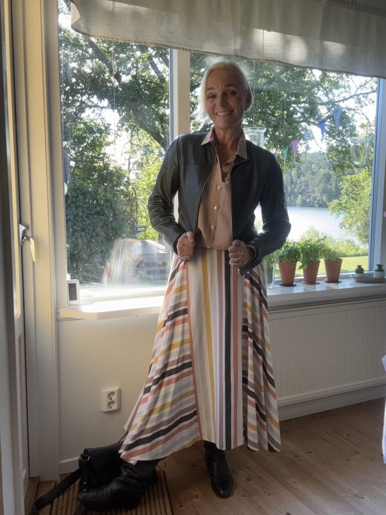 En leende kvinna står inomhus vid ett fönster, klädd i en svart jacka över en ljus blus och en lång, färgglad randig kjol. Hon poserar med händerna i jackan, med solljus och grönska synliga utanför.