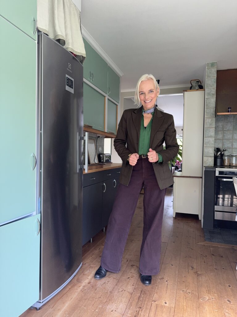 En äldre kvinna med vitt hår står leende i ett modernt kök, klädd i grön skjorta, brun kavaj, vida bruna byxor, svarta skor och en blå halsduk knuten runt halsen. Köket har gröna skåp och trägolv.