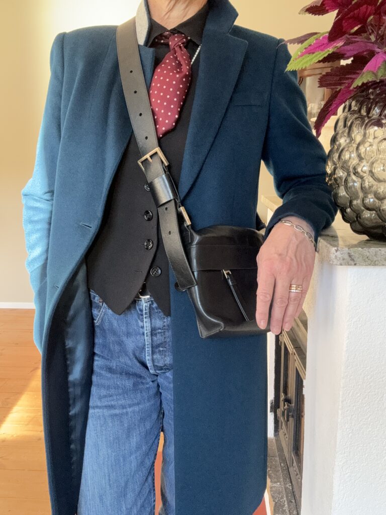 En person klädd i en blågrön överrock, svart väst, röd prickig slips, blå jeans och en svart crossbody-väska står inomhus bredvid en växt i en strukturerad silverkruka. Endast överkroppen och en del av ena armen syns.