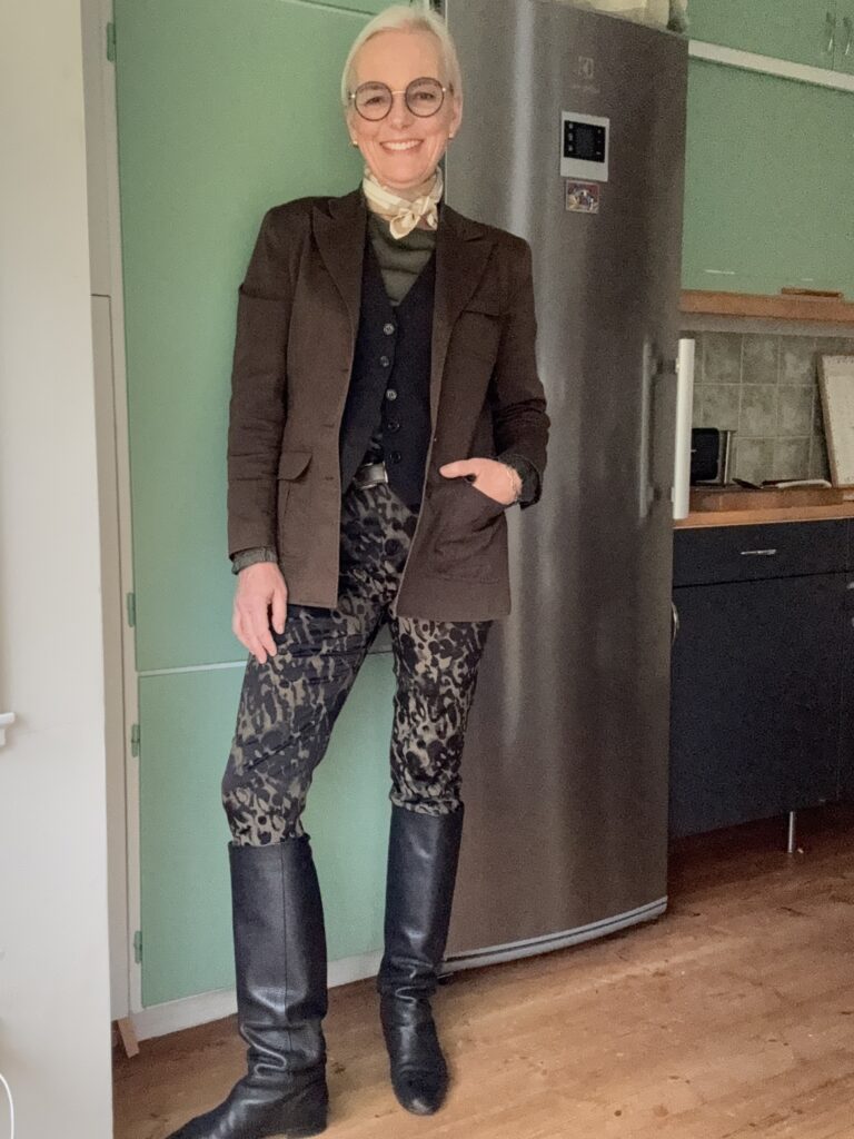 En leende äldre kvinna med kort grått hår och runda glasögon står inomhus. Hon bär en brun kavaj, svart väst, mönstrade byxor, höga svarta stövlar och en guldfärgad scarf och poserar självsäkert framför ett kylskåp i rostfritt stål.