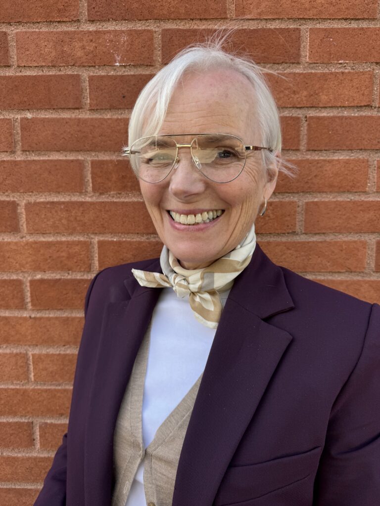 En äldre person med kort vitt hår ler medan han står mot en röd tegelvägg. Hen bär glasögon, mörk kavaj, beige väst, vit skjorta och en ljusmönstrad scarf knuten i nacken.