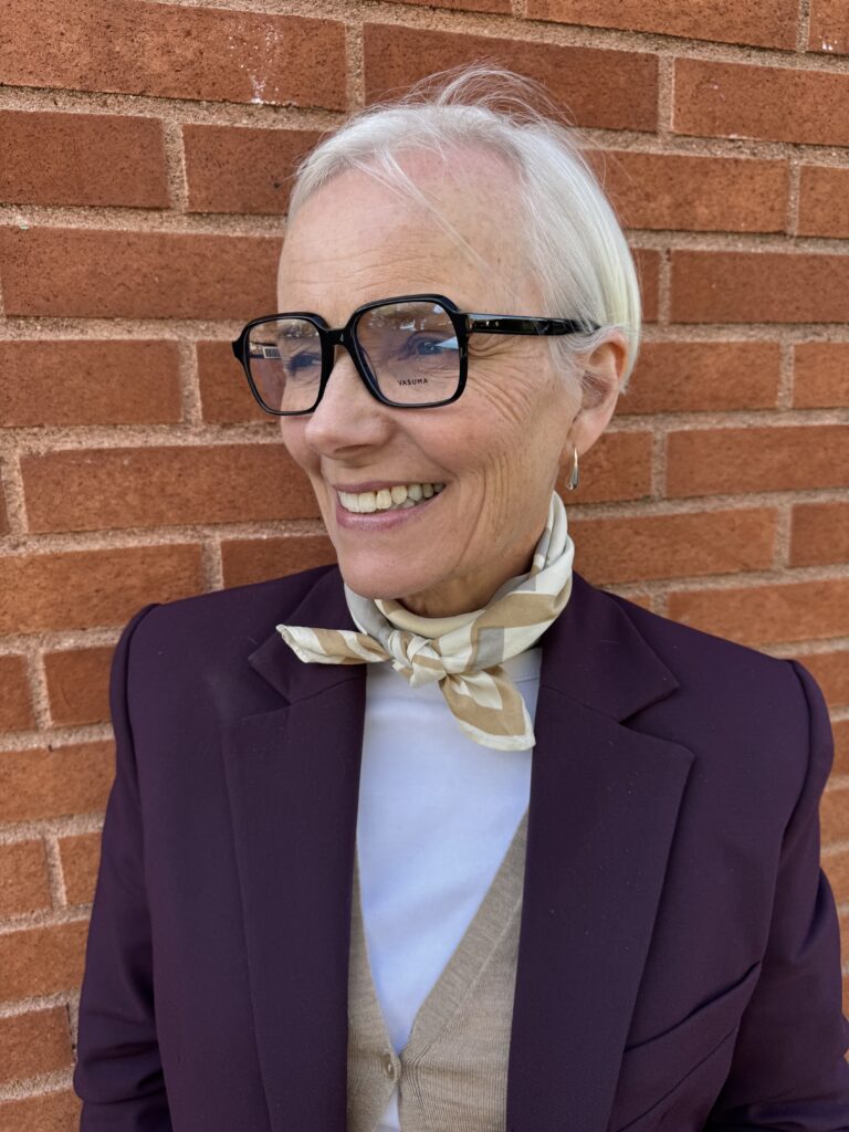 Leende äldre kvinna med kort vitt hår och stora glasögon, klädd i lila kavaj, beige väst, vit topp och en ljusmönstrad scarf, står framför en röd tegelvägg.