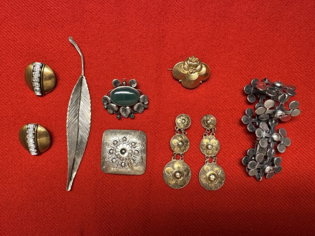 En samling vintagesmycken, bl.a. örhängen, broscher och en fjädernål, på en ljusröd tygbakgrund. Föremålen är guld- och silverfärgade med intrikata mönster och ädelstenar.