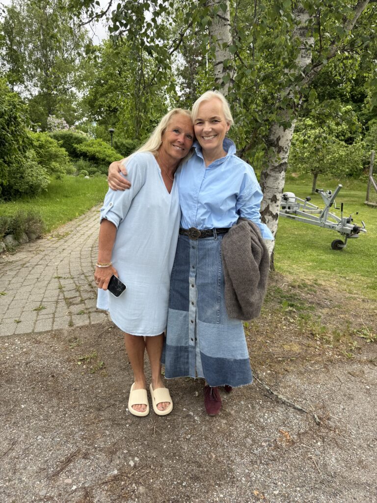Två leende kvinnor står utomhus på en stig, omgiven av grönska. Den ena bär en ljusblå klänning och sandaler, den andra en blå skjorta med en lång lapptäckt jeanskjol och håller i en jacka. De omfamnar varandra och verkar lyckliga.