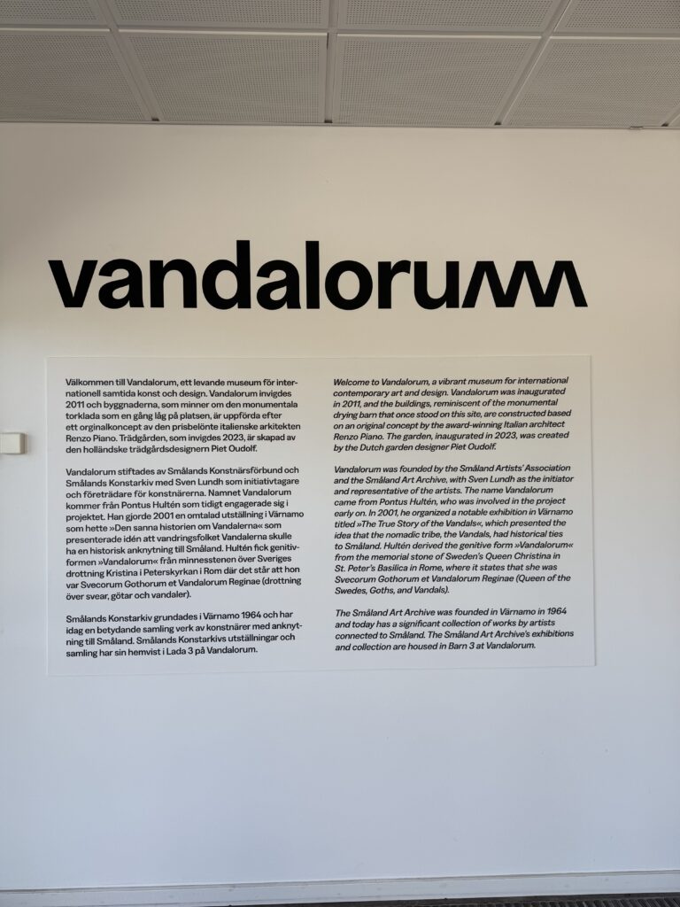 Väggdisplay på Vandalorum museum med svart text "vandalorum" och en skylt på svenska och engelska som beskriver museets grundande, placering i Värnamo och dess inriktning på internationell konst och design.