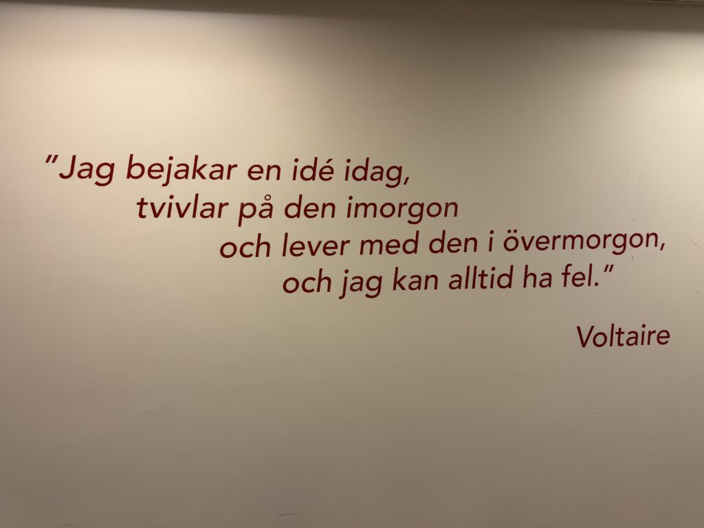Ett citat av Voltaire på svenska står skrivet med röda bokstäver på en beige vägg. Citatet handlar om att omfamna en idé idag, tvivla på den imorgon, leva med den dagen efter och att alltid kunna ha fel.