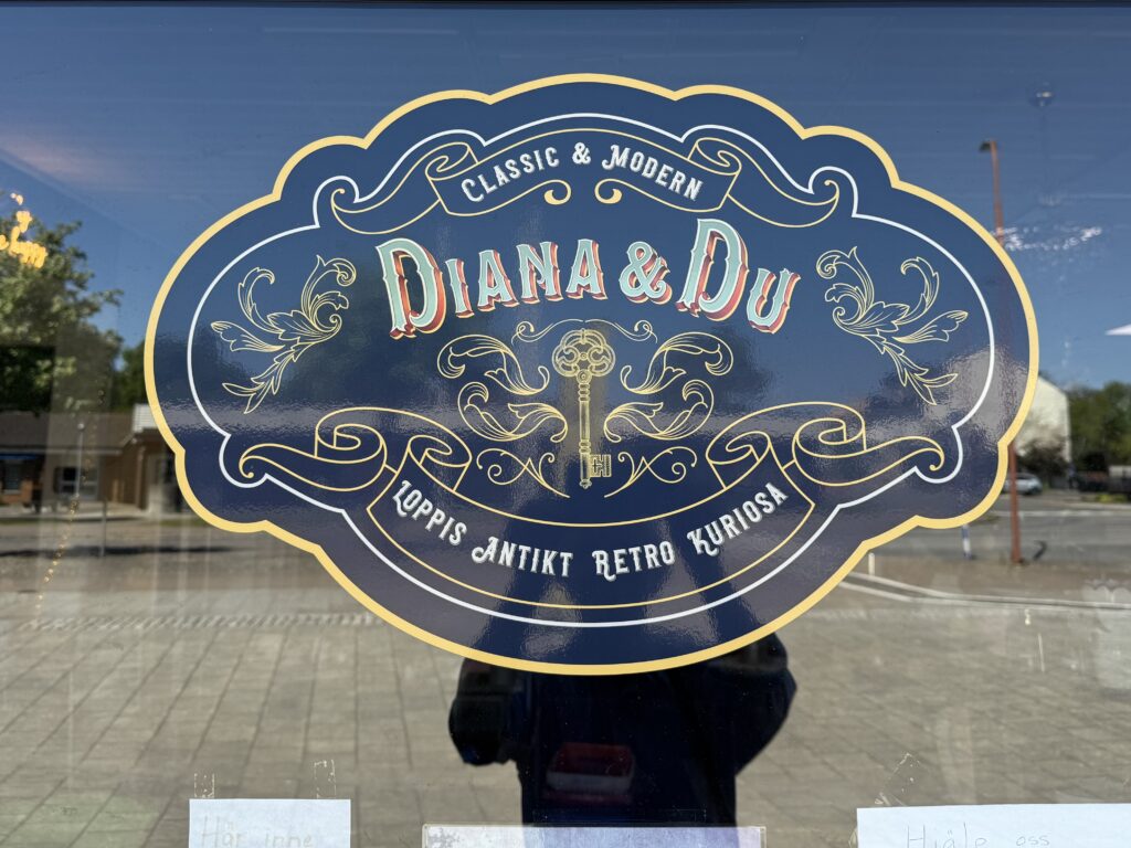 En dekorativ skylt på ett glas har texten "Classic & Modern Diana & Du Loppis Antikt Retro Kuriosa" med guldbokstäver och motiv i vintagestil, bl.a. en nyckel, mot en suddig bakgrund av en gata.
