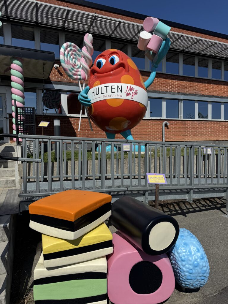 En stor, färgglad staty med godismotiv som håller i klubbor står framför en tegelbyggnad, omgiven av överdimensionerade lakrits- och godisskulpturer en solig dag.
