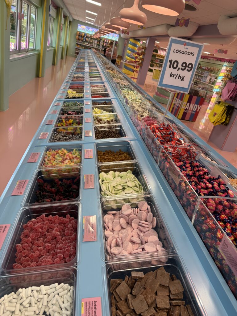 En lång rad färgglada godishinkar står uppradade i en ljus, pastellfärgad butikslokal. Varje låda innehåller olika sorters lösgodis och en skylt visar kilopriset i svenska kronor.