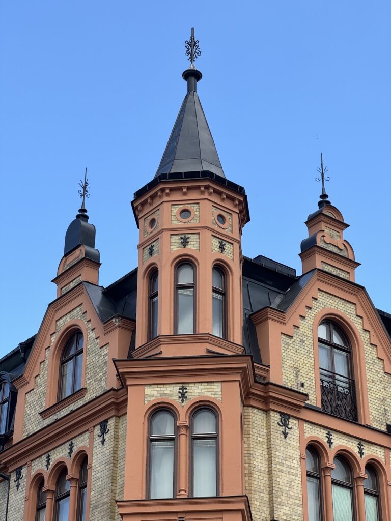 En dekorativ, persikofärgad byggnad med höga välvda fönster och utsmyckade svarta järndetaljer, med ett centralt torn med en spetsig spira och två mindre spiror, mot en klarblå himmel.