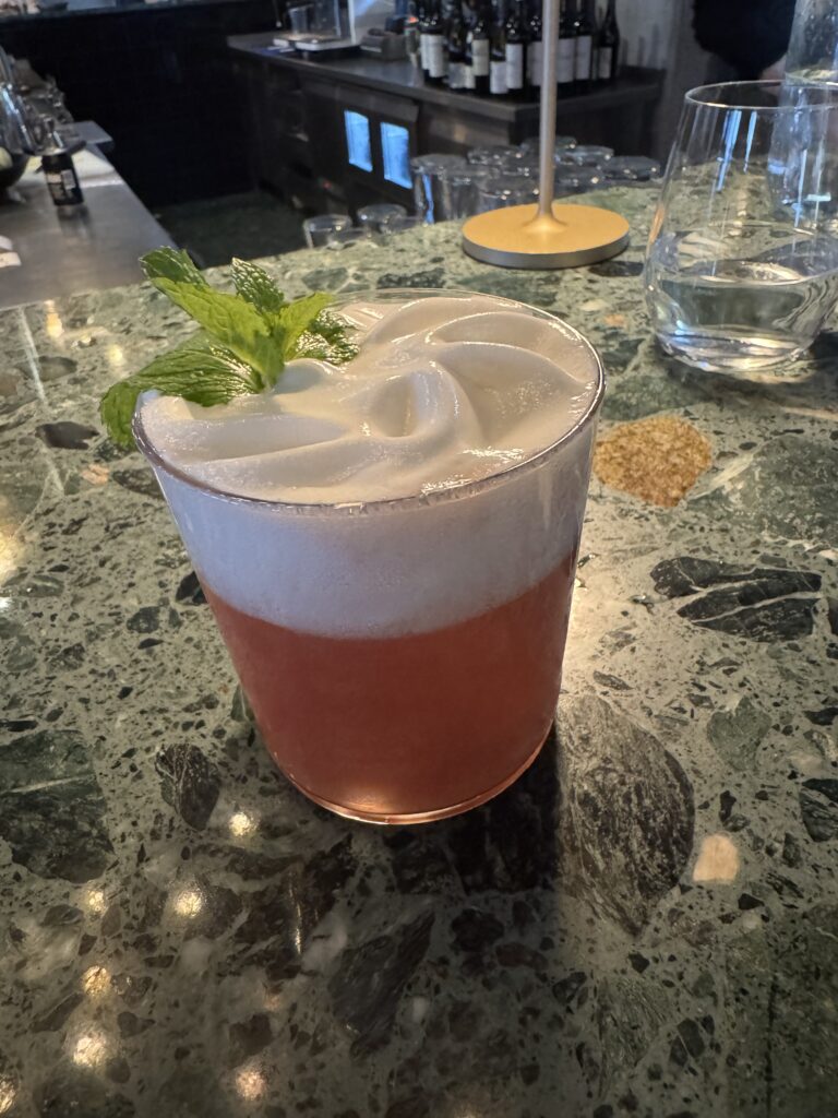 En rosa cocktail med ett tjockt, vitt skum på toppen, garnerad med en kvist mynta, står på en bardisk i grön marmor bredvid ett glas vatten och flaskor i bakgrunden.