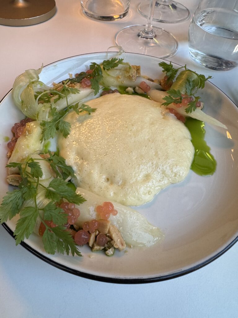 En upplagd gourmeträtt har en krämig, skummande gul sås i mitten, omgiven av örter, vita grönsaksskivor, rosa pärlor, nötter och grön olja, serverad på en vit tallrik med svart kant.