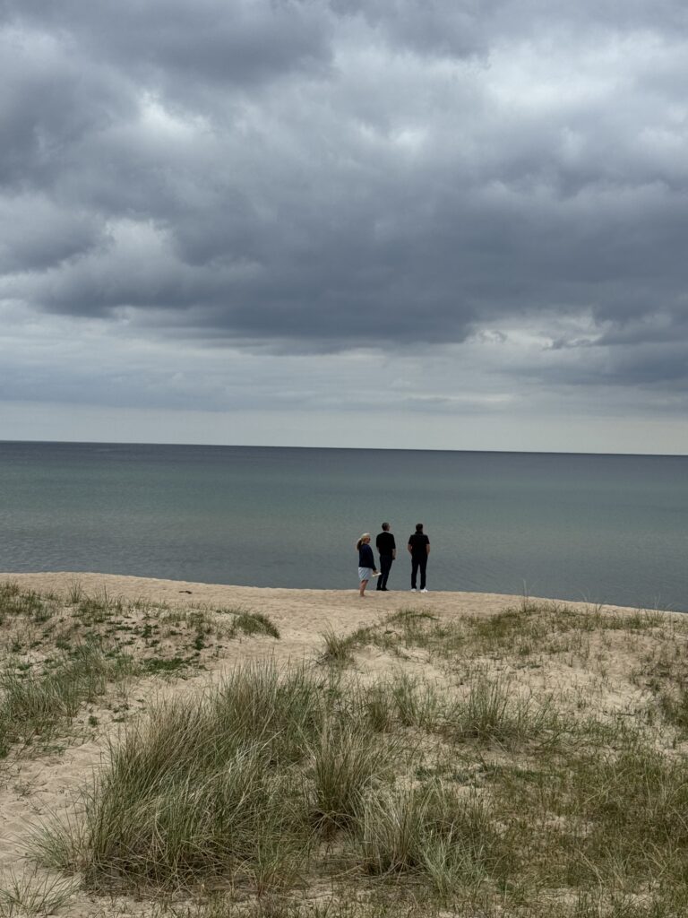 Tre personer står på en sandstrand nära gräsbevuxna sanddyner och tittar ut över en lugn, vidsträckt sjö under en molnig, mulen himmel.