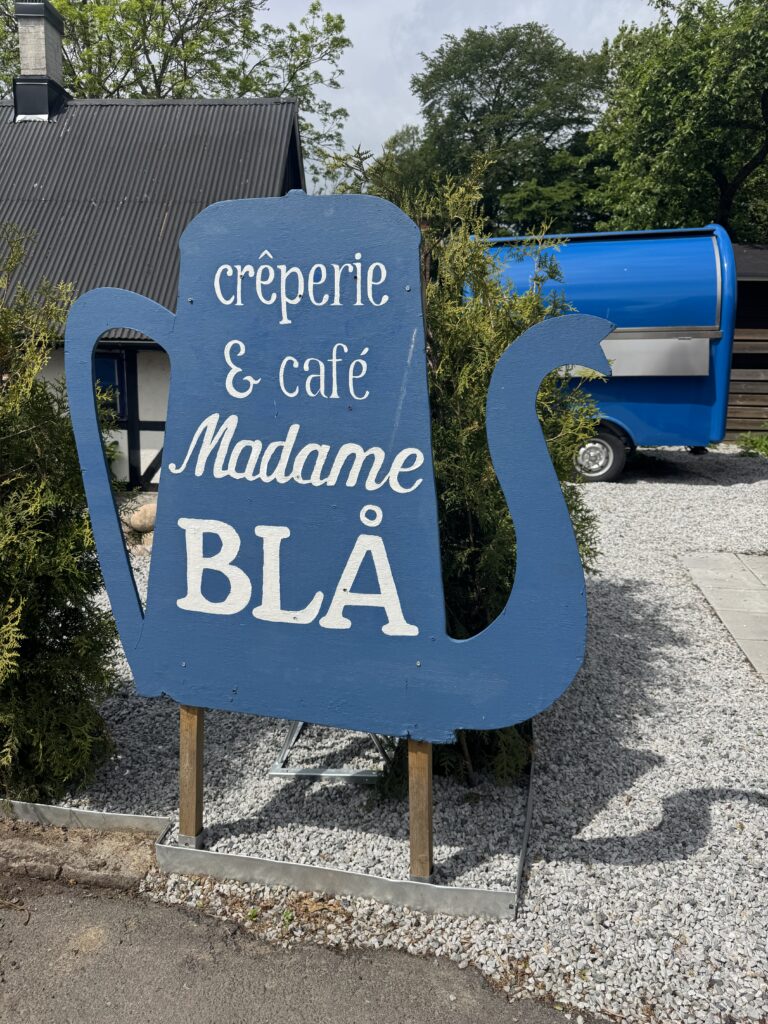 På en stor blå tekannaformad skylt står det "crêperie & café Madame BLÅ" med vita bokstäver. Den står på en grusplan med grönska och en ljusblå husvagn i bakgrunden.