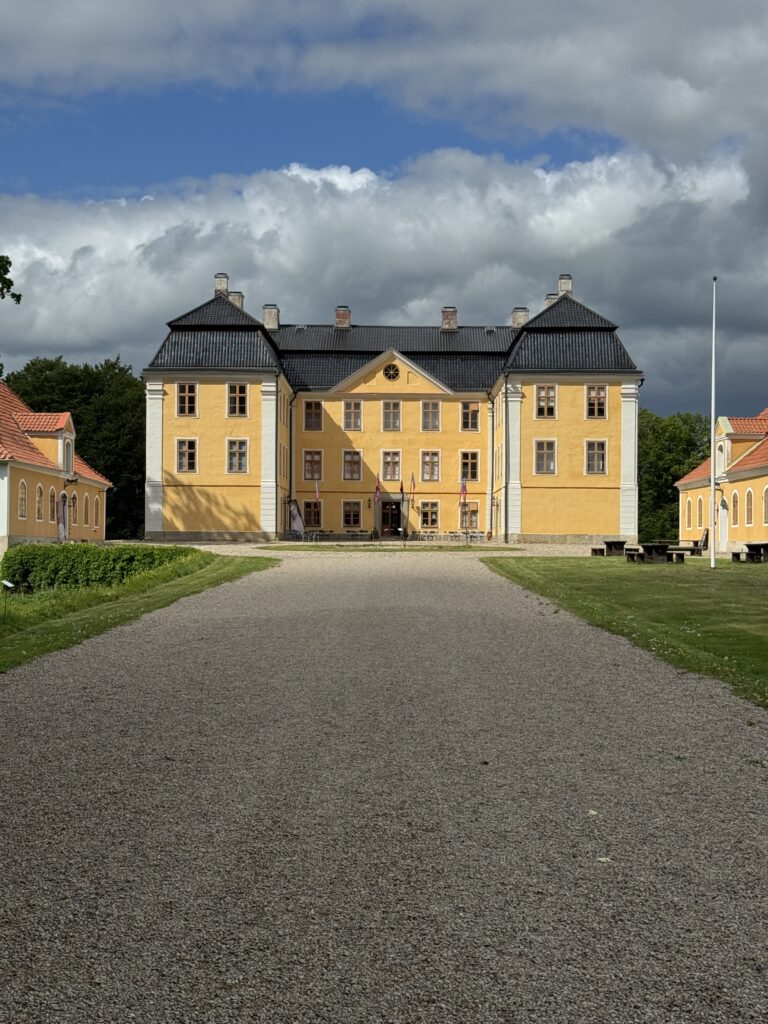 En bred grusväg leder fram till en stor, gul historisk byggnad med svart tak, flankerad av matchande mindre byggnader, under en delvis molnig himmel.