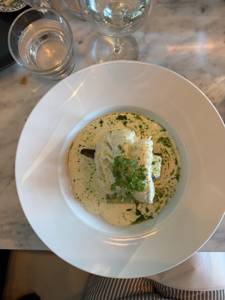 En vit tallrik med en bit vit fisk på grönsaker, garnerad med gröna örter och serverad i en krämig örtsås. Ett vattenglas och ett vinglas står synliga på en marmorskiva.