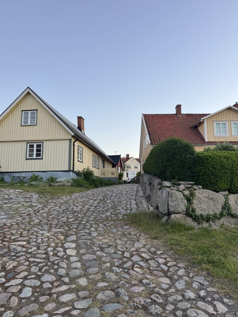 En kullerstensbelagd stig leder mellan två traditionella trähus med lutande tak, ett gräddfärgat och ett gult, under en klarblå himmel. Buskar och stenar kantar stigen på höger sida.