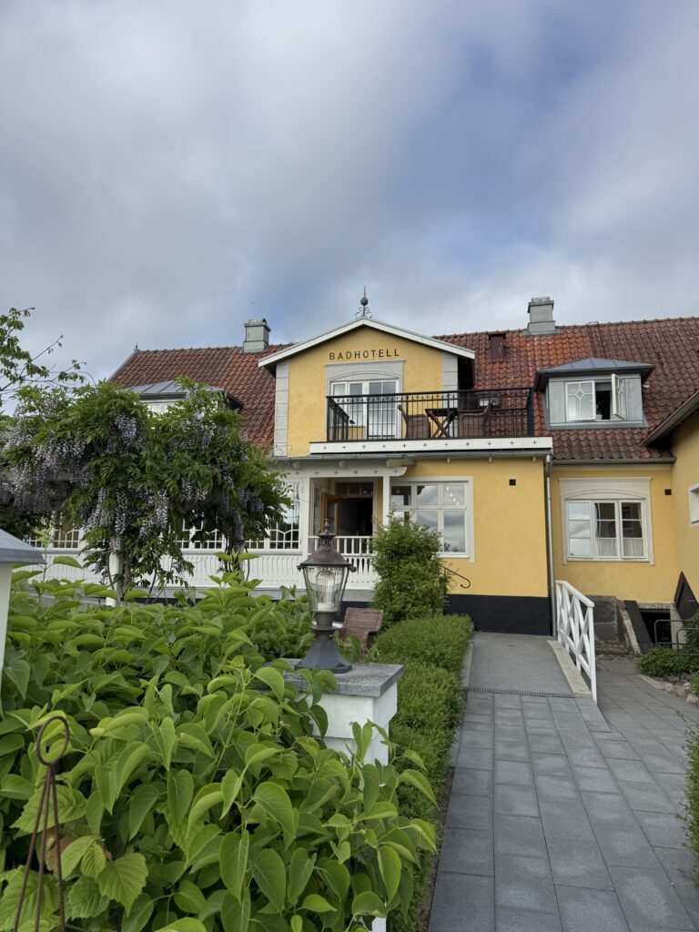 En charmig gul byggnad med rött tegeltak och vita lister, märkt "Badhotell", omgiven av grönska och en asfalterad gångväg som leder till entrén under en molnig himmel.