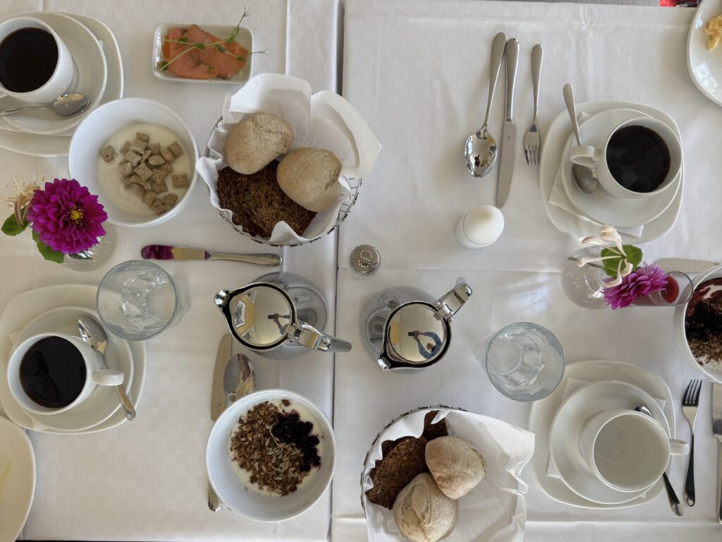 Ett frukostbord dukat för fyra med vita tallrikar, kaffekoppar, korgar med frallor, skålar med flingor och bär, vattenglas och färska blommor på en vit duk.