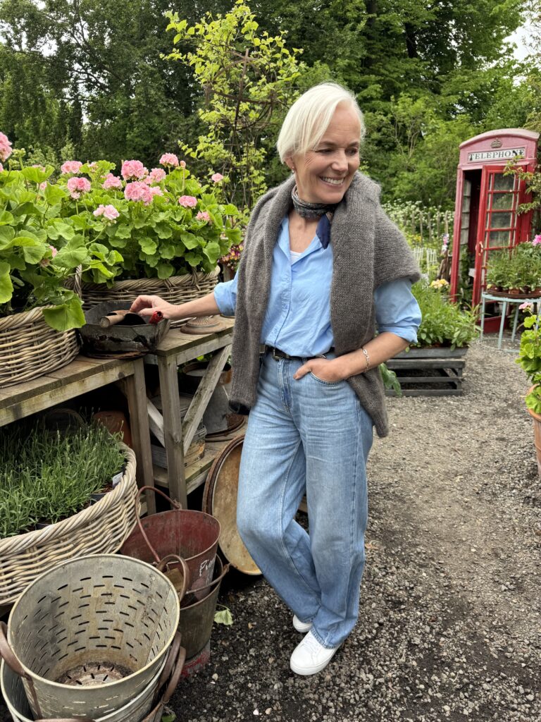 En leende äldre kvinna med kort vitt hår står i ett trädgårdscenter, klädd i ljusblå skjorta, grå tröja över axlarna, blå jeans och vita sneakers, omgiven av krukväxter och korgar.