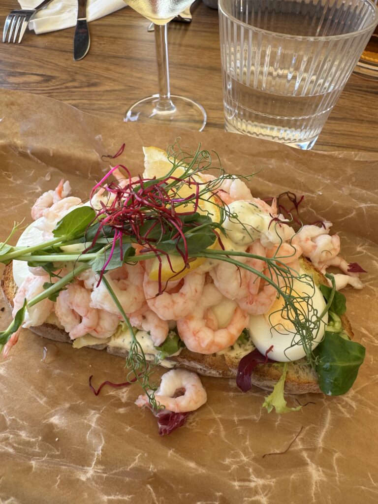 Smörgås med räkor, skivade kokta ägg, blandade grönsaker, citronklyfta, rosa groddar och krämig dressing, serverad på brunt pergamentpapper. Ett glas vatten och ett vinglas finns i bakgrunden.