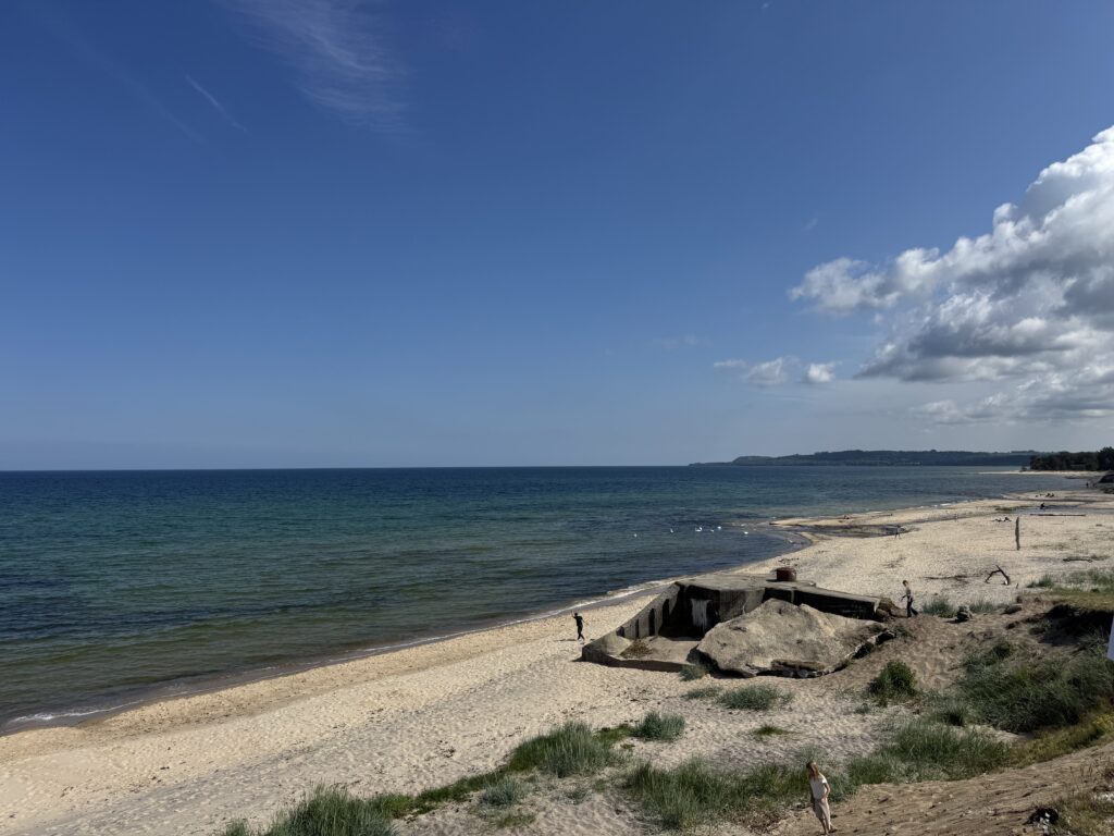 En sandstrand med klarblått vatten, en delvis molnig himmel och några människor som går nära en stor betongkonstruktion. Lite gräs och sanddyner syns i förgrunden.