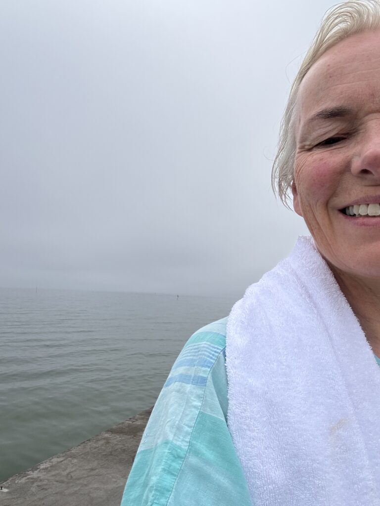 En kvinna med ljust hår och en vit handduk över axeln ler mot kameran; hon befinner sig utomhus vid ett lugnt, grått vattendrag under en mulen himmel. Endast den högra halvan av hennes ansikte och överkropp syns.