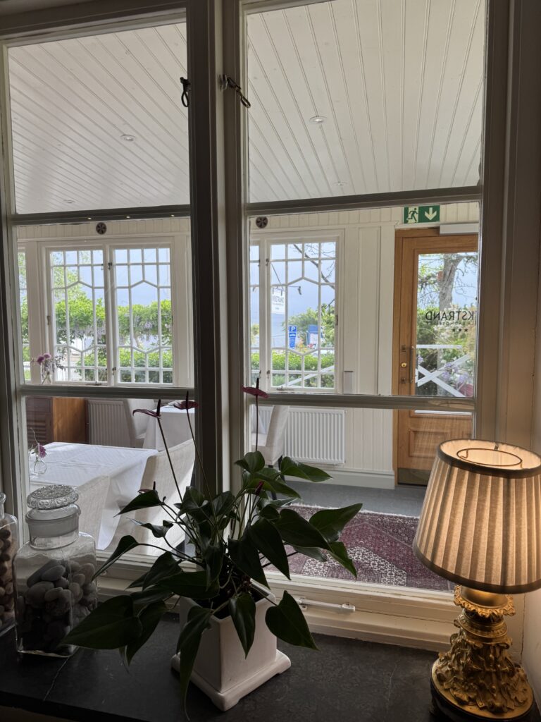 Vy genom ett fönster in i ett ljust, mysigt rum med vita väggar och möbler, en krukväxt på fönsterbrädan, en tänd bordslampa, glasburkar och en trädörr som leder ut till grönska.
