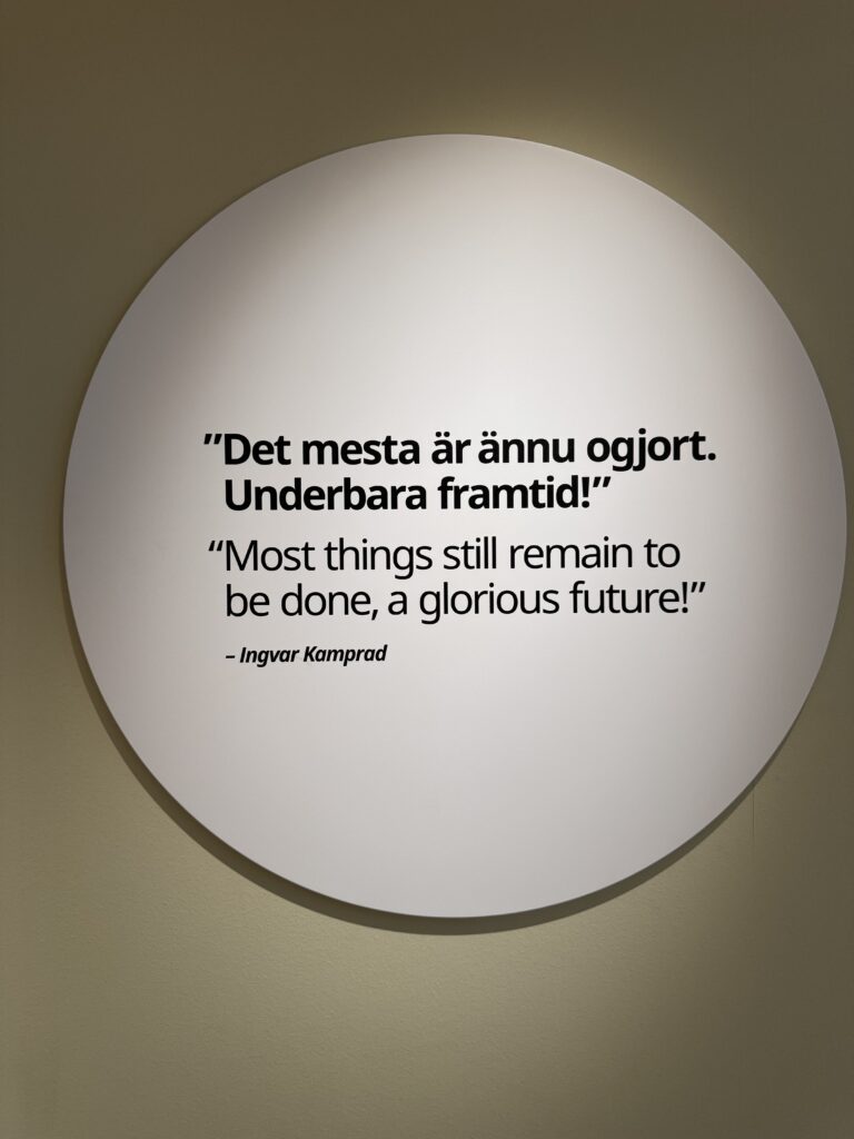 En vit rund skylt på en beige vägg visar ett citat på svenska och engelska: "'Det mesta återstår att göra, en härlig framtid!' - Ingvar Kamprad.".