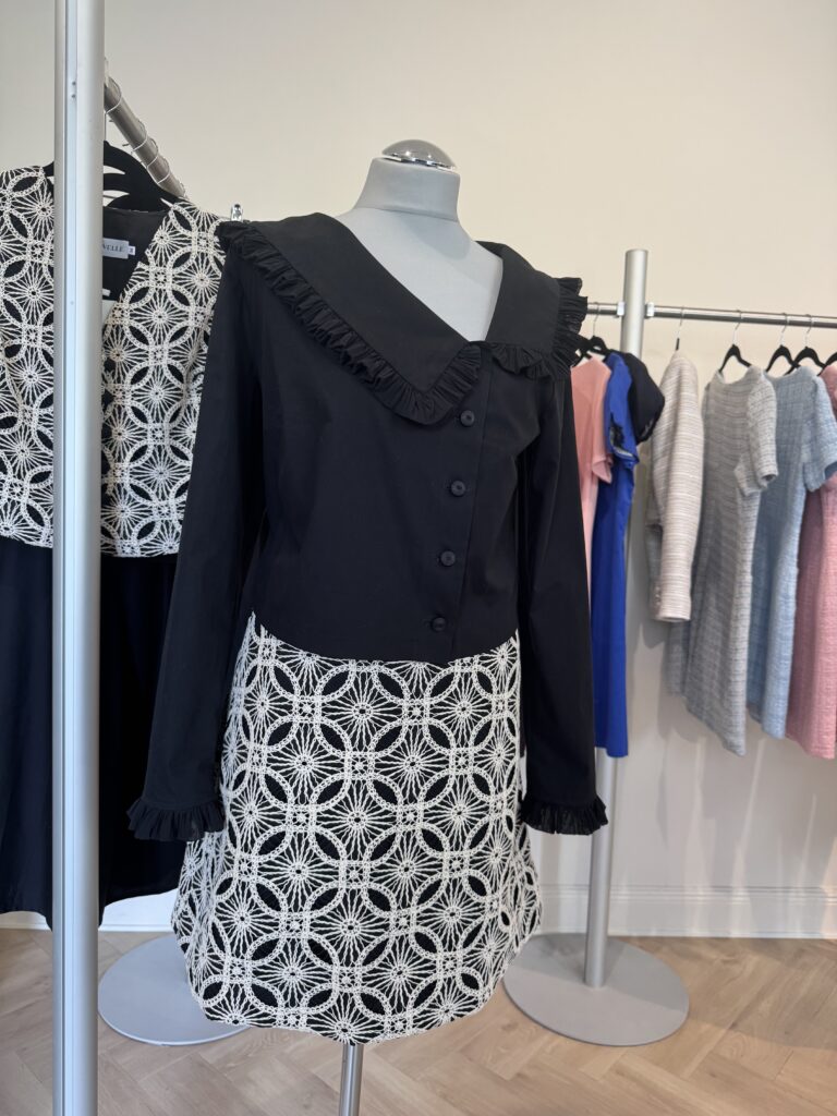 En skyltdocka visar en klänning med svart, långärmad topp med volangkrage och manschetter samt vit kjol med geometriskt svart blommönster. Andra färgglada plagg hänger på ställningar i bakgrunden.