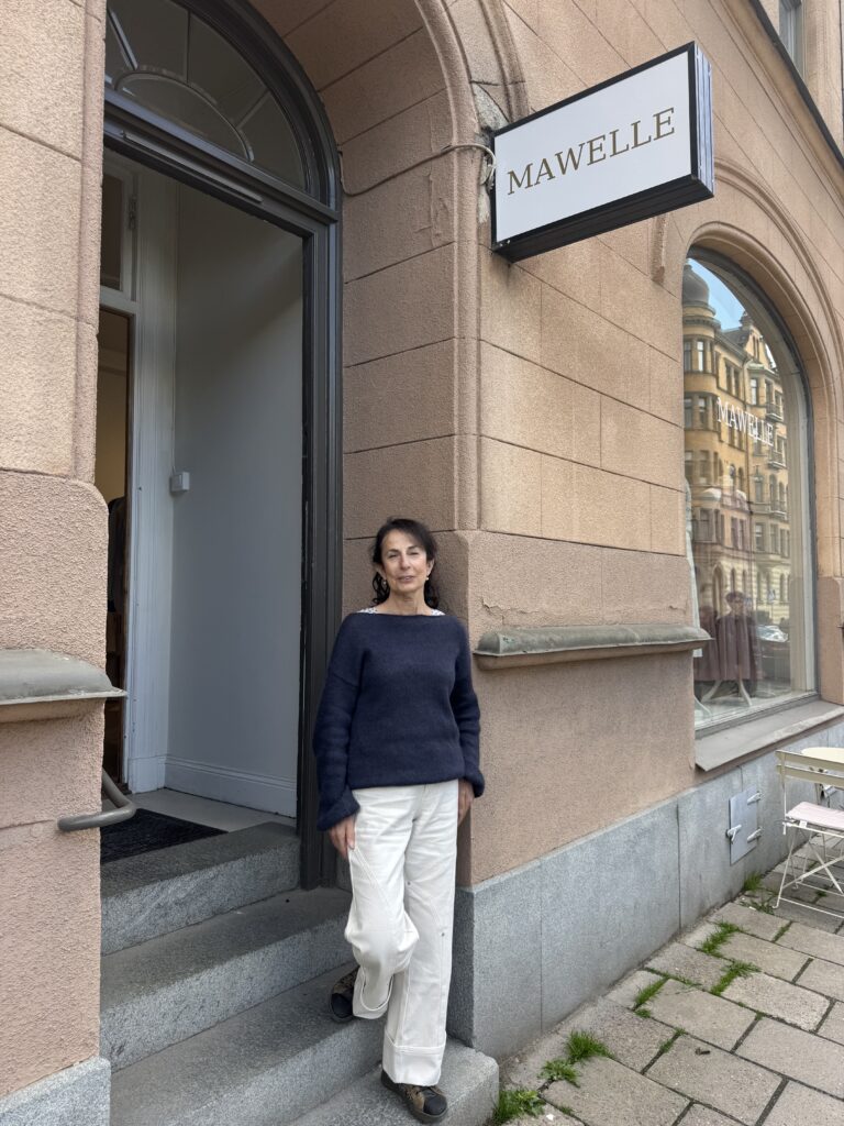 En kvinna i marinblå tröja och vita byxor står på trappan utanför en byggnad med en skylt där det står "MAWELLE". Byggnaden har stora fönster och solbrända stenväggar. En stol och ett bord syns på trottoaren.