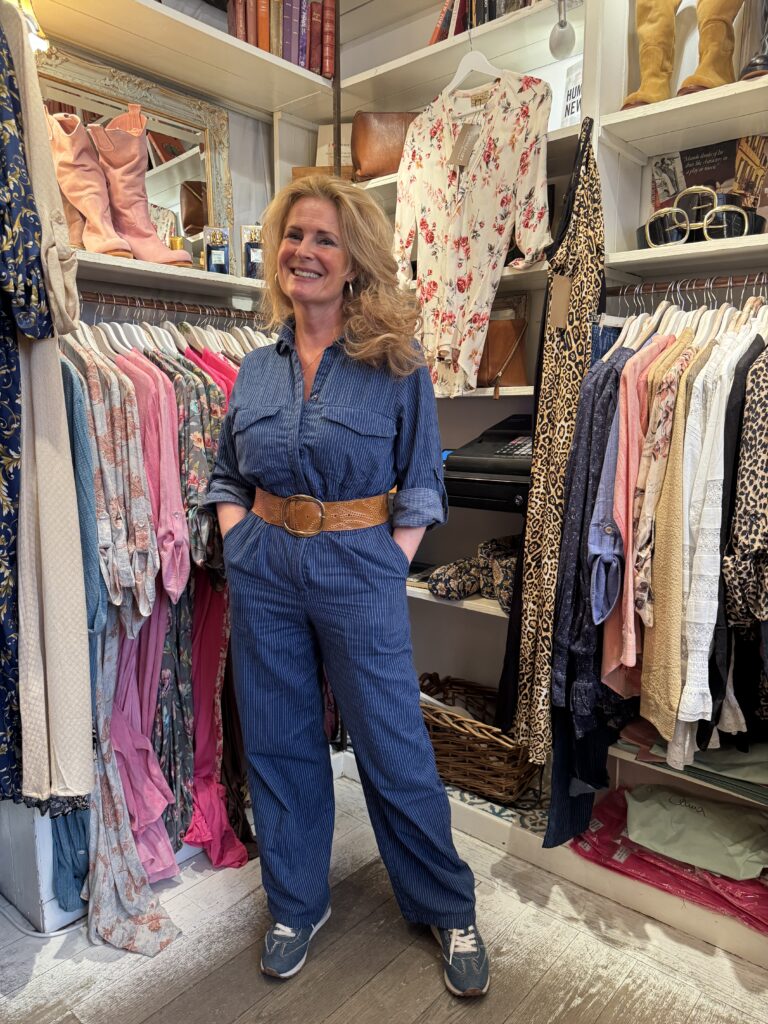 En kvinna med vågigt blont hår, klädd i en blå jumpsuit, brunt bälte och sneakers, står leende i en klädbutik omgiven av färgglada kläder, skor och accessoarer på hyllor och ställningar.