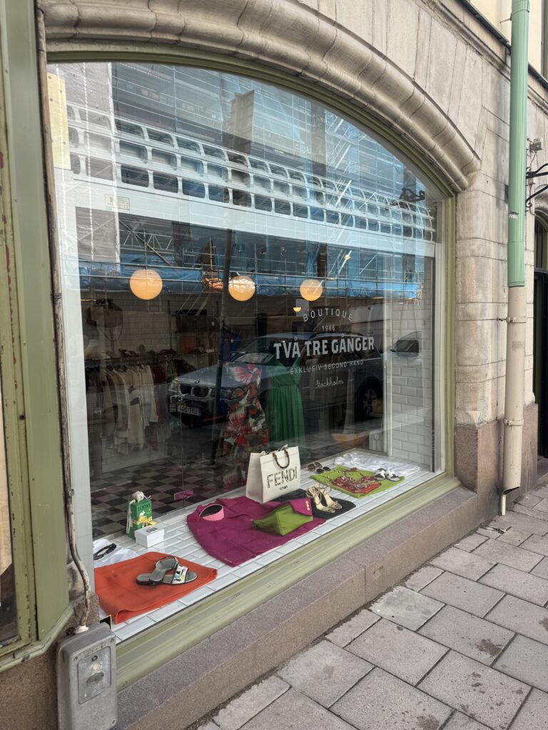 En klädbutik med stora välvda fönster visar färgglada kläder och accessoarer, bland annat en Fendi-väska, på trottoarsidan. Butikens interiör är synlig med hängande plagg och runda lampor.