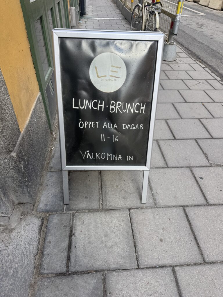 På en trottoarskylt utanför en byggnad står det: "LUNCH BRUNCH, ÖPPET ALLA DAGAR 11-16, VÄLKOMNA IN", en inbjudan att komma in på lunch eller brunch varje dag kl. 11-16. En cykel är parkerad i närheten.
