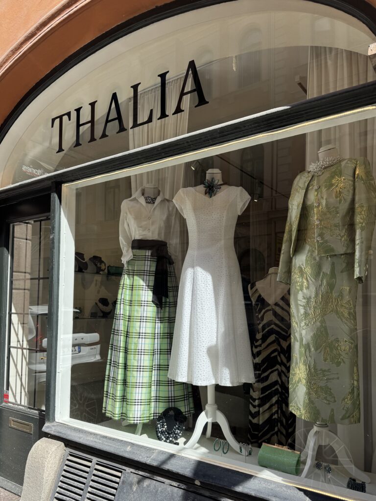 Skyltfönster på "THALIA" med tre skyltdockor i eleganta klänningar: en vit topp med grönrutig kjol, en vit kortärmad klänning och en grönblommig jacka över en matchande klänning. Solen skiner på skyltfönstret.