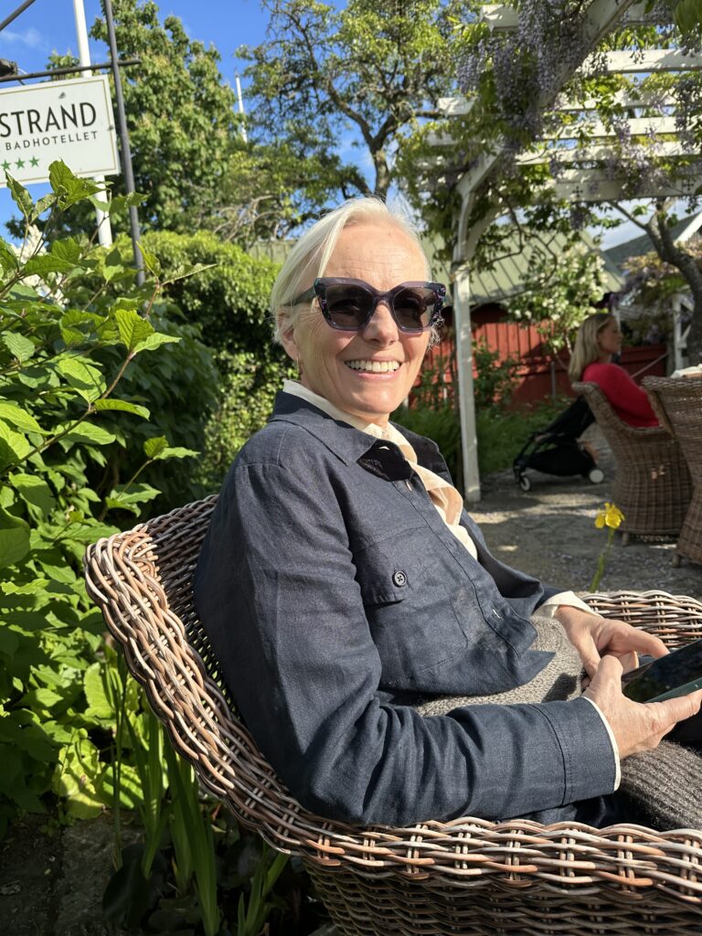 En äldre kvinna med kort blont hår och solglasögon ler när hon sitter i en korgstol utomhus en solig dag och håller i en telefon. Gröna växter och en hotellskylt syns i bakgrunden.