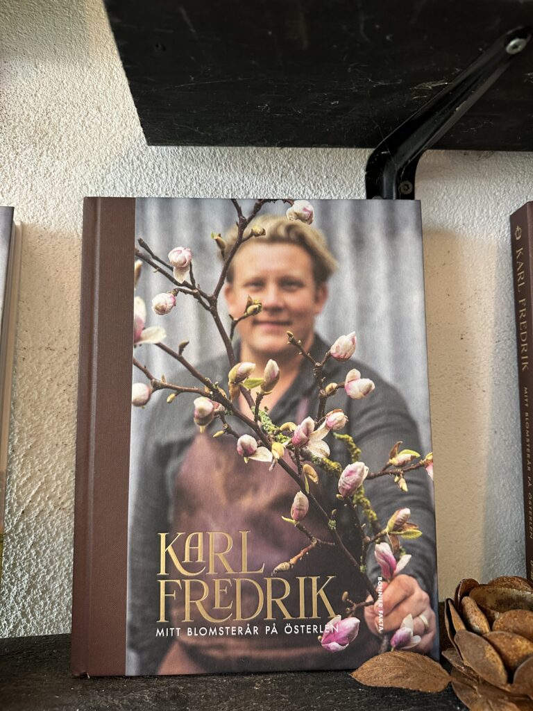 En bok med titeln "Karl Fredrik: Mitt blomsterår på Österlen" står upprätt på en hylla. Omslaget visar en person som håller i en gren med rosa och vita blommor mot en neutral bakgrund.
