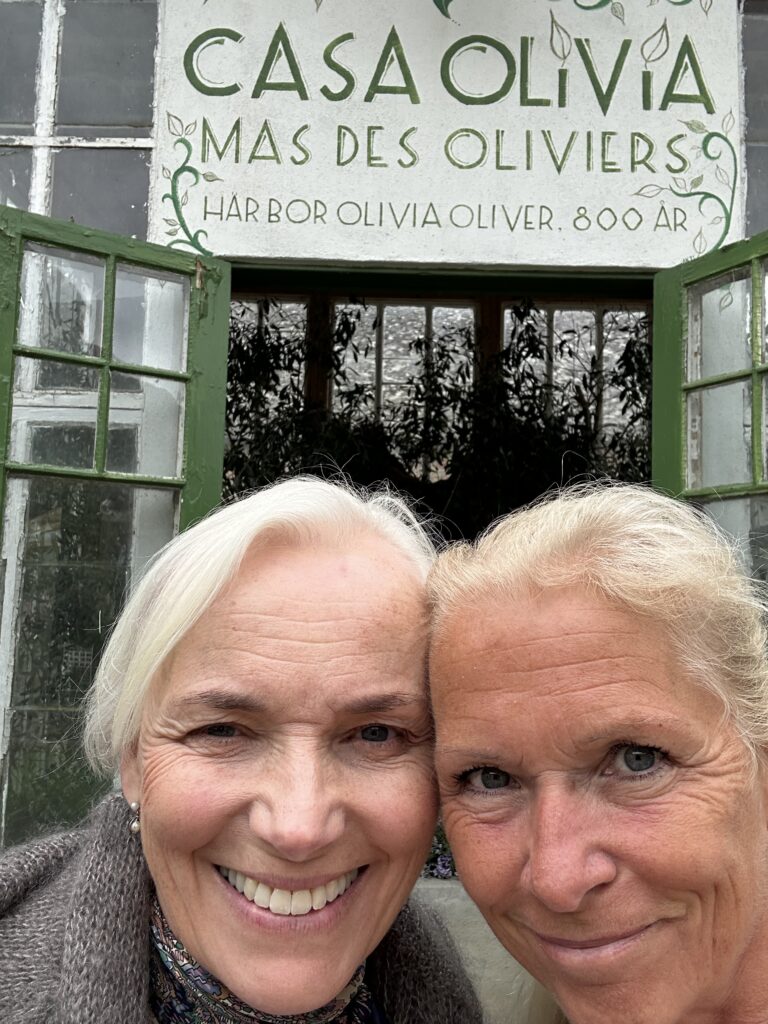 Två leende kvinnor med ljust hår tar en närbildsselfie framför en byggnad med grönmålade fönster och en skylt ovanför med texten "Casa Olivia Mas des Oliviers" och text på svenska.