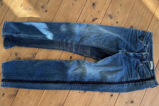 Ett par blåjeans ligger platt på ett trägolv och visar tecken på slitage och blekning, särskilt på lår och knän. Solljuset skapar högdagrar och skuggor på både jeansen och golvet.