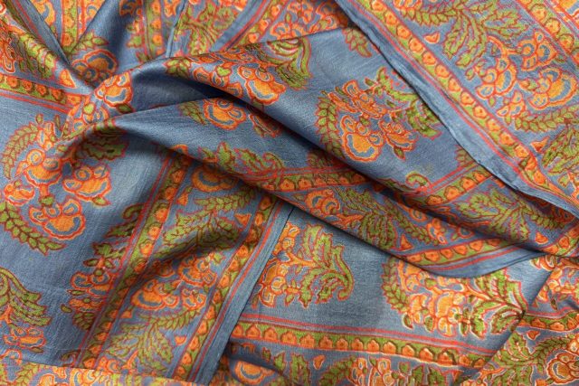 Närbild av blått tyg med intrikata orange och gröna paisley- och blommönster, vikt med synliga veck och dekorativa kanter.