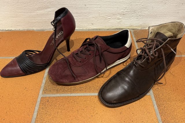 Tre olika skor på ett klinkergolv: en vinröd högklackad sko, en rödbrun sneaker med vita detaljer och en brun snörkänga i läder.