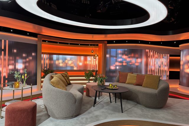 En elegant TV-studio med böjda grå soffor, färgglada kuddar, små bord med blommor och ljus samt en orangevit och rund lampa i taket. Bakgrunden har abstrakta orange och vita ljusmönster.