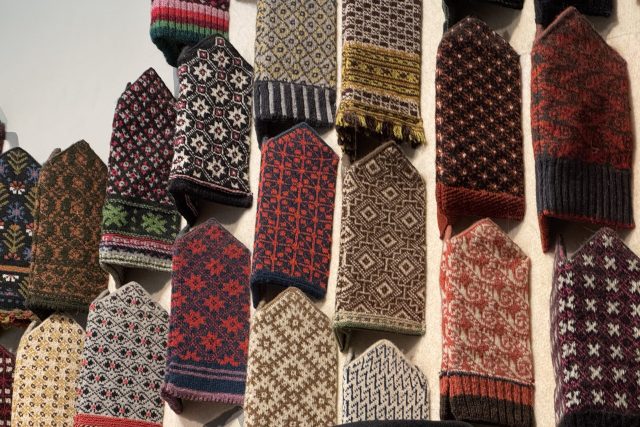 En uppvisning av färgglada, intrikat mönstrade stickade vantar i vertikala rader på en vägg, som visar olika traditionella mönster och motiv.