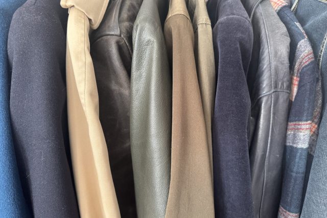 Närbild på flera jackor och rockar i olika färger och tyger, bl.a. beige, grön, svart läder, marinblå och rutigt mönster, som hänger sida vid sida på en ställning.